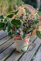 Foraged Christams decorations: IIlex - holly, Hedera - Ivy, Papaver - poppy seed heads, Viburnum and a cinnamon stick in a terracotta pot pained with mistletoe and a red raffia bow.