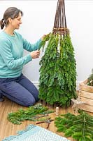 Woman using brown garden twine to attach pieces of conifer - juniperus foliage to willow obelisk frame