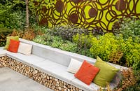 Large sawn Yorkstone stone bench with colourful cushions and log storage underneath. The Sunken Retreat. RHS Malvern Spring Festival, 2016. Design: Ann Walker for Graduate Gardeners
