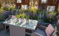 Outdoor acrylic rattan dining furniture with table and chairs with orange cushions on patio surrounded by a lucent copper slate raised bed with colourful planting of yellow and purple flowering plants, ornamental grasses with rusted corten steel screens. The RNIB Community Garden. RHS Hampton Court Flower Show July 2018  - Designer: Steve Dimmock and Paula Holland - Sponsor RNIB, Magus Private Wealth 