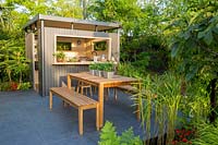 An outdoor entertaining space with metal outdoor kitchen bar set on porcelain stone patio with wooden bench and table. B and Q Bursting Busy Lizzie Garden