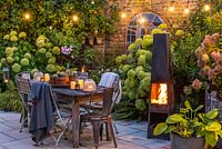 A dining area in a courtyard garden lit by a string of bulbs and candles, heated by a modern outdoor chimenea. Planting behin includes Hydrangea 'Limelight', Rosa 'Blush Noisette', Anemone hybrida 'Elfin Swan' and ferns.