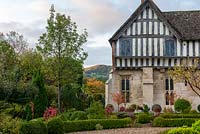 A glimpse of Cooper's hill beside the facade of a part timbered, Mediaeval manor house overlooking a formal box parterre.