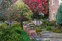 View along an old terrace, beyond a mature ornamental cherry with autumn's red foliage. Urns planted with begonias and periwinkle.