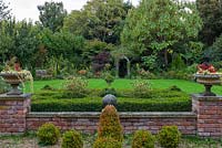 View from a terrace over a small clipped box parterre flanked by urns planted with begonias and periwinkle, over a lawn to a rose arch flanked by borders of maples, rudbeckias, hydrangeas and dahlias, below an Indian bean tree.