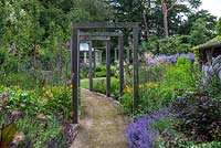 A contemporary lower garden with a path leading beneath a pergola made up of eight standalone oak frames that frame views over the borders of Aconites, Daylilies, Dahlias, Geum, Sea hollies, Catmint, Campanulas, Salvias and Verbenas.