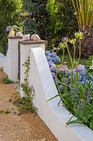 Rendered white wall at the edge of a garden with mixed borders with plants including Agapanthus and Hydrangea. The Dream of The Indianos, Hampton Court Flower Show, 2019.