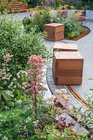 View through public space with moveable wooden cube seating on a rail, mixed summer borders edged with rill water feature. The Crest Nicholson Livewell Garden - Hampton Court Flower Show 2019 