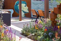 View through hot summer borders towards contemporary living and dining area and outdoor kitchen with female mural on wall in the background. The Lower Barn Farm Outdoor Living Garden - Hampton Court Flower Festival 2019 