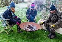 People wrapped in warm clothes, roasting marshmallows over a fire in a Corten Steel fire pit on a frosty winter day. 