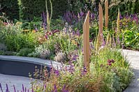 Compacted path sprialing around beds full of colourful summer perennials with inscribed wooden posts. The Cancer Research UK Pledge Pathway to Progress - Hampton Court Flower Festival, 2019.