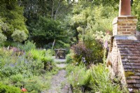 Path through a lush cottage garden leading to a simple decked dining area with planting including ferns and lots of self seeders including foxgloves and aquilegias in a cottage garden in June