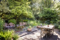 Simple decked dining area surrounded by dense trees and shrubs in a cottage garden in June. Four contemporary orange metal seats are arranged in a row to one side with planting including Carex testacea and blue festuca.