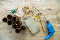 Drill, hammer, nails, pliers, scissors, pencil, ruler, wooden board, garden wire, sandpaper, rope, string and tin cans laid out on a wooden surface