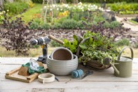 Drill, Wooden board, masking tape, pencil, scissors, rope, pots, trowel, watering can, gloves, goggles and basket of plants laid out on a wooden surface