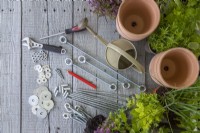 Steel rods, tensioners, hooks, connectors, nuts, washers, screwdriver, spanner, watering can, pencil, terracotta pots, plants and compost laid out on a wooden surface