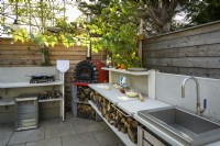 Outdoor kitchen with red pizza oven, metal sink, hob and wood storage in small modern family garden