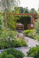 The Viking Friluftsliv Garden. An  outdoor dining and kitchen area is seen over Pinus mugo 'Mops' and beds of gaura, penstemon, coneflowers and astrantias, beneath tall birches.