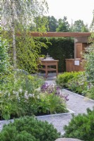 An outdoor dining and kitchen area is seen over Pinus mugo 'Mops' and beds of gaura, penstemon, coneflowers and astrantias, beneath tall birches.