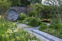 A Place to Meet Again. Hampton Court Flower Festival 2021. An urban courtyard is planted in restful tones of grey, green and white, with height added by a pergola draped in star jasmine. A circular water feature on the wall upcycles pipes and taps.