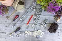 Steel rods, hooks, connectors, nuts, washers, screwdriver, spanner, watering can, pencil, terracotta pots, plants and compost laid out on a wooden surface