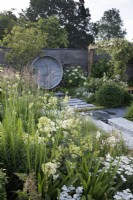 In A place to Meet Again Garden, the path paved with smooth dutch style pavers cuts through the planting which uses a green and white palette with Agapanthus and Hydrangea paniculata; at the back, a water feature of repurposed brass taps stands against a wall of stacked concrete slabs- Designer: Mike Long - Sponsors: Association of Professional Landscapers, Kebur Garden Materials, Creepers Nurseries, Landscape Plus.