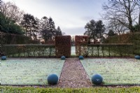 View  through the frosty lawns to to the focal point at the end of the garden, as the sun rises. Painted balls form manmade structure alongside the clipped hornbeam and box hedges.