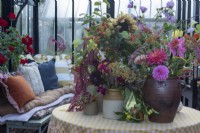 A table in the greenhouse that has a collection of vases of cut flowers, including: dahlias, sunflowers, cosmos,sedums and rose hips.