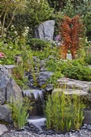 View over waterfall, with planting such as Typha laxmannii among rocks, to rusty metal sculpture. Bodmin Jail: 60Ã‚Â° East - A Garden Between Continents, RHS Chelsea Flower Show 2021