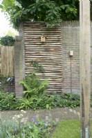 Roll of chestnut paling hung over brick wall with bird house attached, ferns and groundcover beneath.