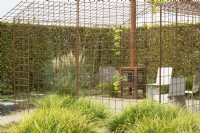 House frame of corten steel grid with corten stove and Miscanthus in border.