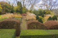 View over mature clipped hedges of Yew in front of wave-form hedge of Beech. Area planted with ornamental grasses in blocks with several clipped Yews in cone form; image taken with drone. November. Autumn.