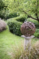 Decorative metal sphere on a stone plinth surrounded by pheasant grass, Anemanthele lessoniana, in a June garden