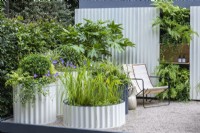 Assorted powder coated, corrugated steel containers are planted with shade-loving plants such as Fatsia japonica, ivies, hostas and periwinkles, whilst vertical planting of ferns contrasts against the corrugated steel wall panels.