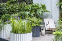Assorted powder coated, corrugated steel containers are planted with shade-loving plants such as Fatsia japonica, ivies, hostas and periwinkles. A wide shallow container makes a miniature water feature, planted with aquatic grasses and pickerel weed.