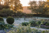 The kitchen garden in late autumn, dusted in frost. Vegetable beds of herbs, leeks and dahlias, with apple step-over cordons beyond.