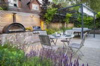 View across the flower bed to dining area with wooden table, chairs, bespoke kitchen unit with integrated pizza oven and bbq grill. 