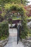 An arch and a wrought iron gate that is half open leads to a seating area with two wrought iron chairs and a table. Whitstone Farm. NGS garden, Devon. Spring. 