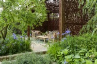 Sitting arrangement in pavillion  of metal screens  with  Morris' Willow Boughs pattern also on cushions, surrounded by herbaceous beds with blue Iris siberica, Rodgersia  and  Salix matsudana tortuosa trees, dragon's claw willow - Morris and Co. Garden