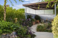 Garden with covered seating area beside a pond planted with Iris 'White Swirl' and Carex grasses with  green, purple, bronze and blue planting including Sambucus  'Black Beauty' and Betula nigra tree in raised bed  - SSAFA Sanctuary Garden. Designer: Amanda Waring