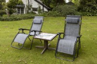 Two reclining garden chairs and a mosaic table on a lawn at Lewis Cottage, NGS Devon garden. Spring.