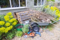 Pallets, compost, compost scoop, watering can, lining, scissors, tiles, screws, screwdriver, pencil, tape measure, drill, basin, twigs, logs and various dried plants laid out on the ground