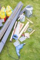 Drill, hammer, saw, spirit level, pencil, tape measure, screws, nails, sandpaper, pliers, wire, paint, paint brush, string, watering can, and wood pieces laid out on the ground