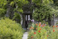 A paved path to an old stone ruin with beds planted with perennials including poppies, Papaver rhoeas. The Garden House, Yelverton, Devon. Summer. 