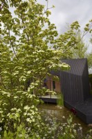 A Garden Sanctuary by Hamptons. A charred Larch cabin set amongst predominantly green and white enclosed planting. Designer: Tony Woods. RHS Chelsea Flower Show 2022. Gold Medal. Summer. May.