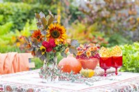 Autumnal table display with Squash, containers of Chrysanthemums, berries and crab-apples with a vase of Beech sprigs, helianthus and Chrysanthemums and a red tableware set