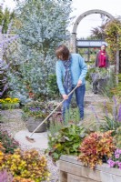 Woman sweeping patio with woman pushing wheelbarrow in background