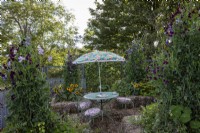 Straw bale seating area with recycled woodchip as ground cover in cottage style garden. Old fashioned printed fabric sun shade and elegant metal chairs
