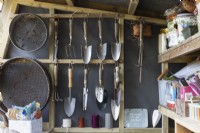 A neat garden shed with trowels and garden sieves hanging at one end and various gardening paraphernalia on shelves on the right hand side. Autumn.