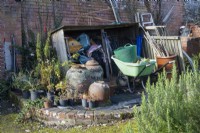A wooden store, set against a red brick wall, holds a variety of plastic sacks with various gardening parahphanlia in front including wheelbarrow, clay cloches, plastic trug and bamboo canes. Regency House, Devon NGS garden. Autumn
